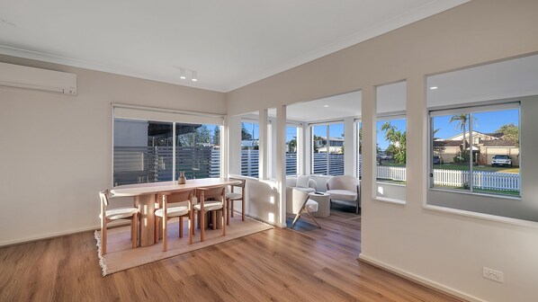 Una habitación amplia con una mesa y sillas de madera, ventanas grandes que ofrecen vistas del barrio y una puerta corrediza de vidrio que da a un espacio exterior.