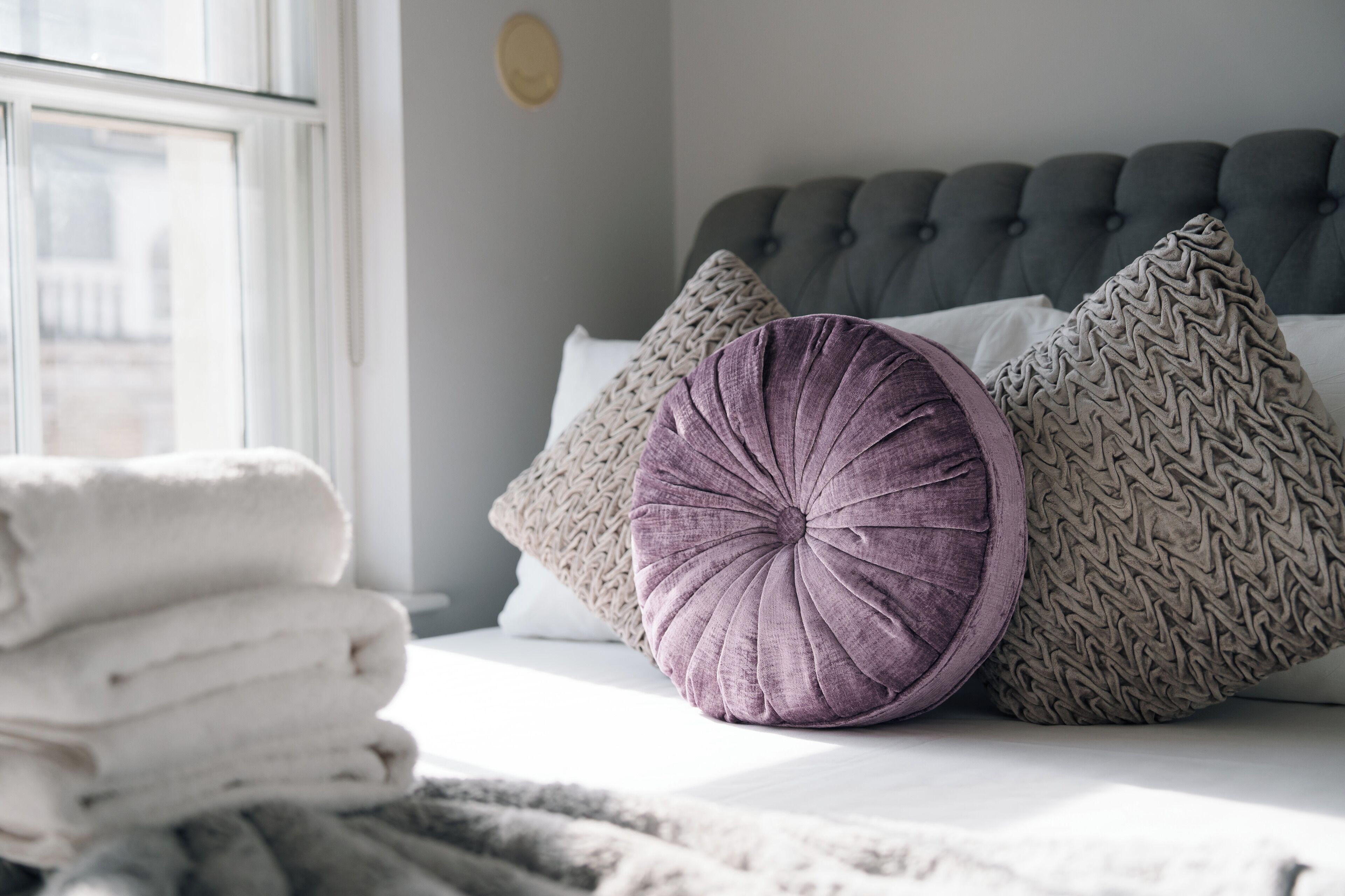 Ein ordentlich bezogenes Bett mit grauem, gepolsterten Kopfteil, einem Stapel weißer Handtücher und zwei dekorativen Kissen.