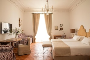 A hotel room with a checkered sofa, two beds, a desk, a chandelier, and a large window with curtains. - Casa Pizarro Hotel (Cáceres)