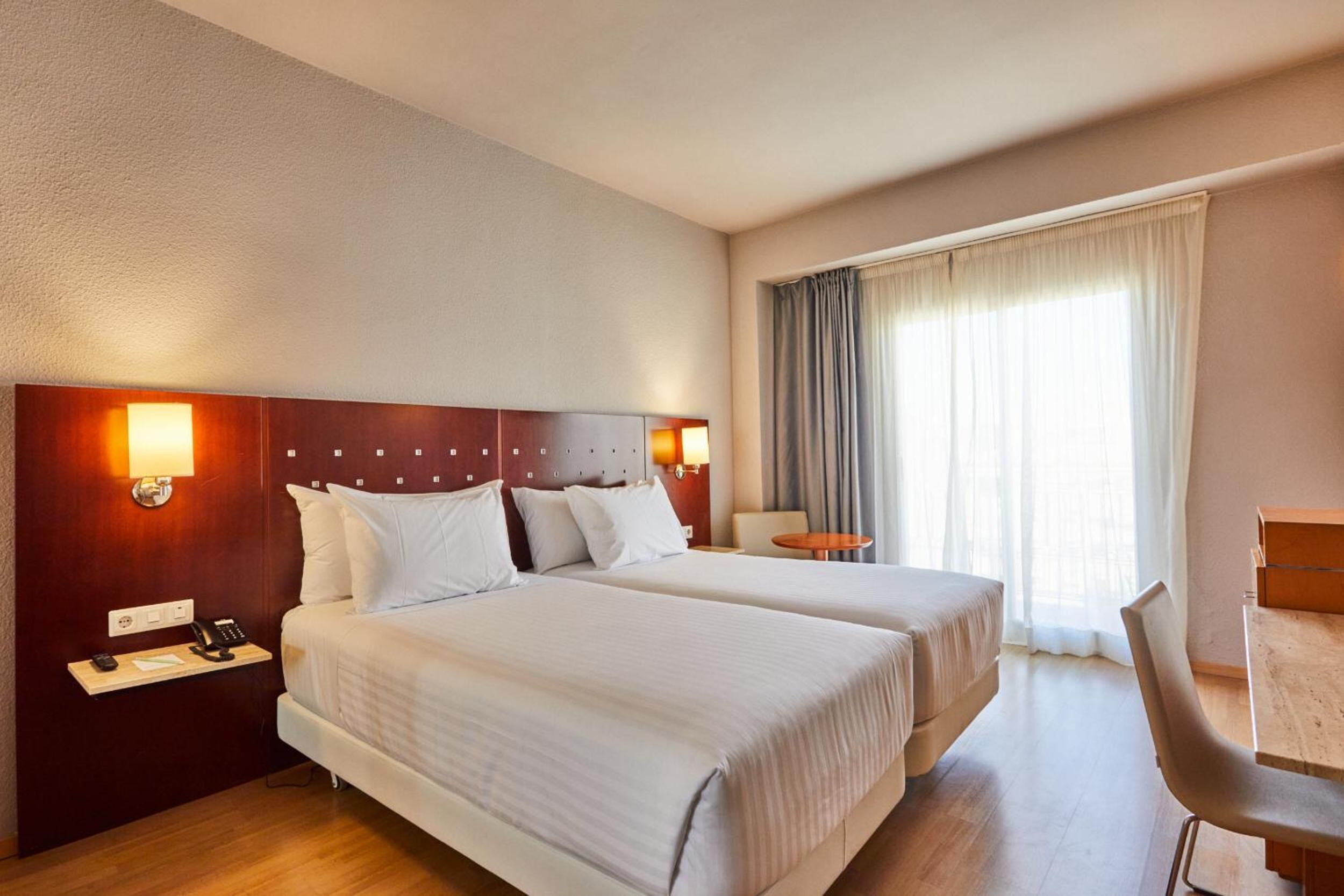 A hotel room with two beds, a desk, a chair, and a window with curtains.