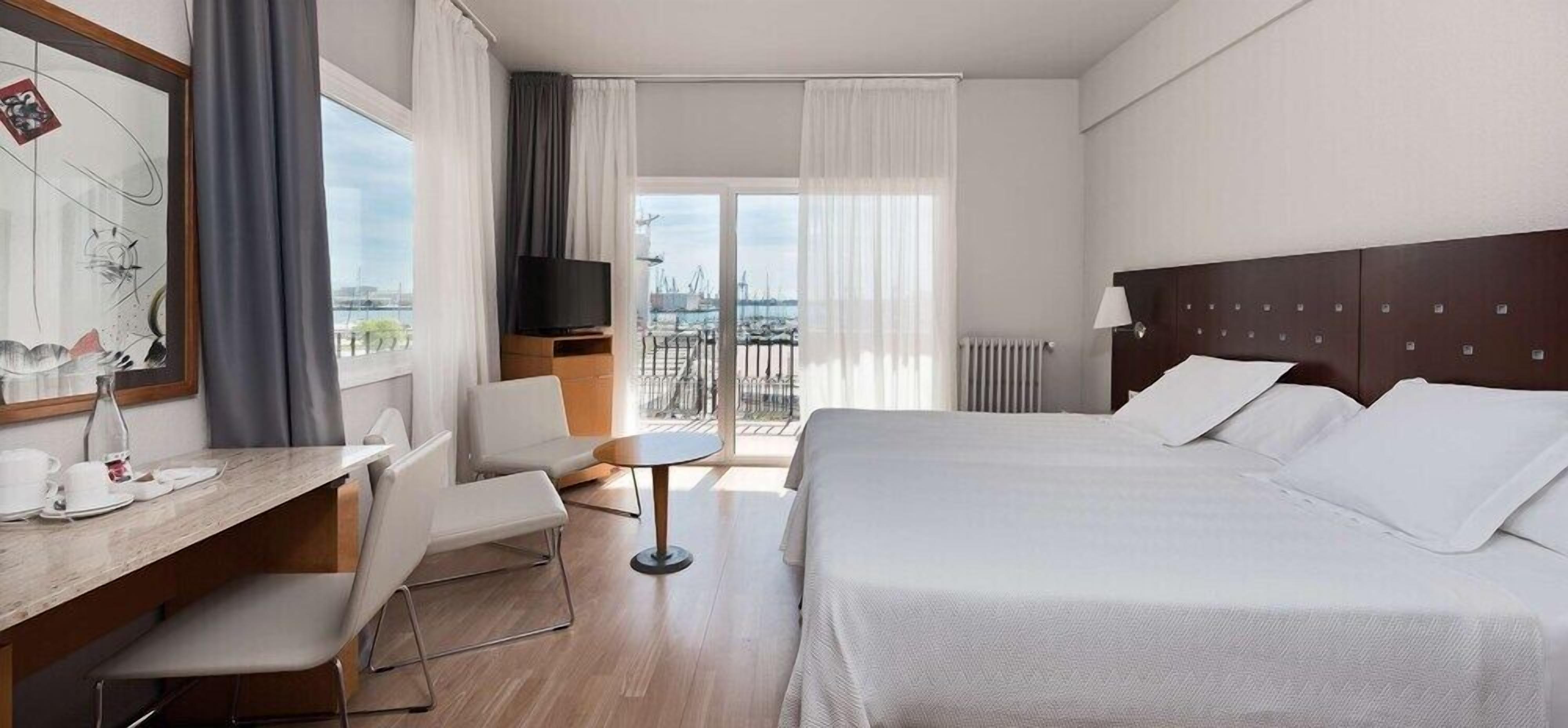 A hotel room with a bed, a desk with a chair, a small table, and a TV.