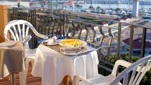Un balcon avec une table dressée pour deux personnes, offrant une vue sur une marina avec des bateaux et sur le lointain paysage urbain.