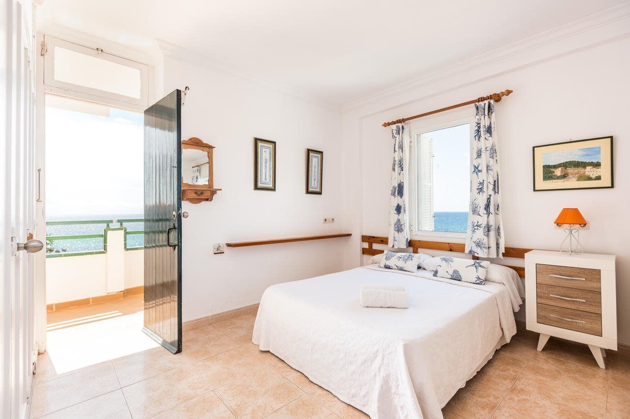 A bedroom with a bed, a nightstand, a window with curtains, a door leading to an outdoor balcony, and a wall-mounted shelf.