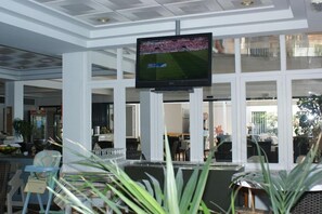 A spacious lobby with a large flat-screen TV mounted on the ceiling, a reception desk, and a seating area with chairs and tables. - Vistamar Apartments (Es Migjorn Gran)