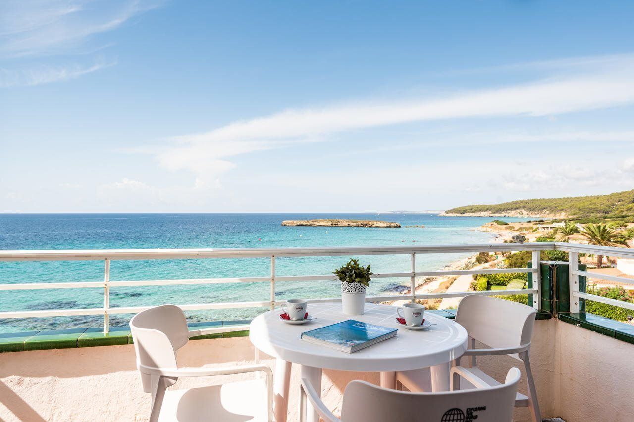 A balcony with a view of the sea, white chairs, a table with a book and cups, and a potted plant.