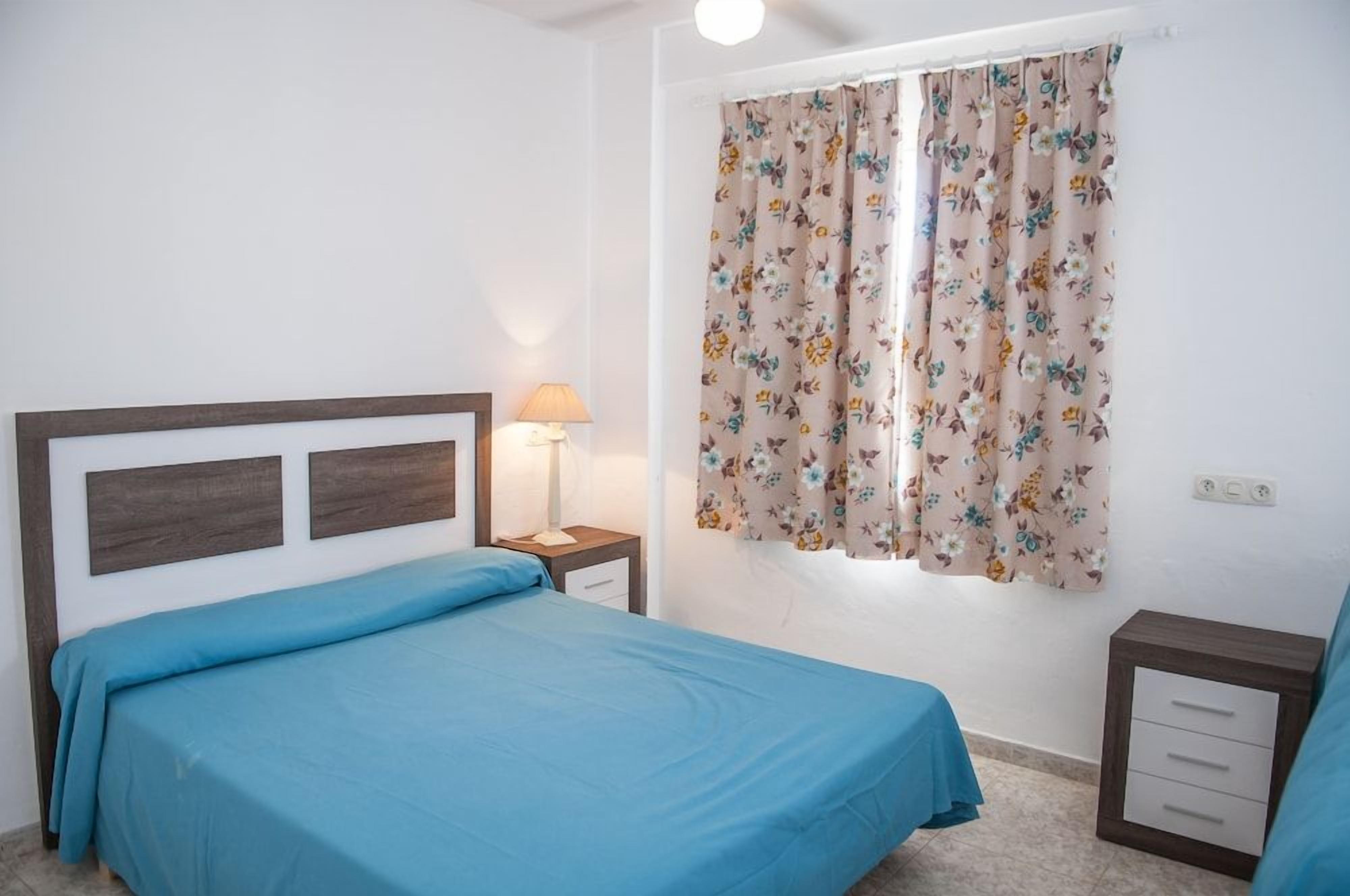 Une chambre à coucher avec un lit, une table de chevet, une lampe et une fenêtre ornée de rideaux à motifs floraux.