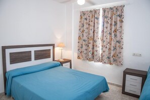 A bedroom with a bed, a nightstand, a lamp, and a window with floral curtains. - Vistamar Apartments (Es Migjorn Gran)