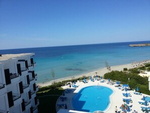 Un área de piscina con sillones de descanso y sombrillas, una playa al fondo y un edificio con balcones.