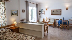 Habitación de hotel acogedora con cama, escritorio, silla y una mesita. Tiene una ventana con cortinas, un estante en la pared con botellas y un cuadro enmarcado.