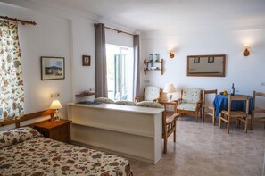 Une chambre d’hôtel confortable, avec un lit, un bureau, une chaise et une petite table. Il y a une fenêtre avec des rideaux, une étagère fixée au mur où sont posées des bouteilles, et un tableau encadré.