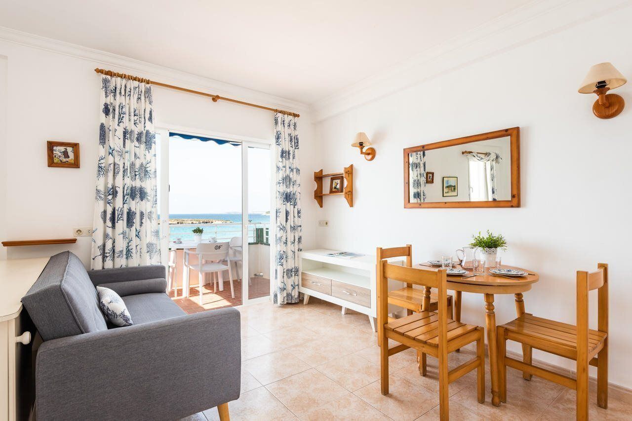Una habitación con sofá, mesa de comedor, sillas, un mueble bajo y vista al mar a través de una puerta.