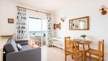 Una habitación con sofá, mesa de comedor, sillas, un mueble bajo y vista al mar a través de una puerta.