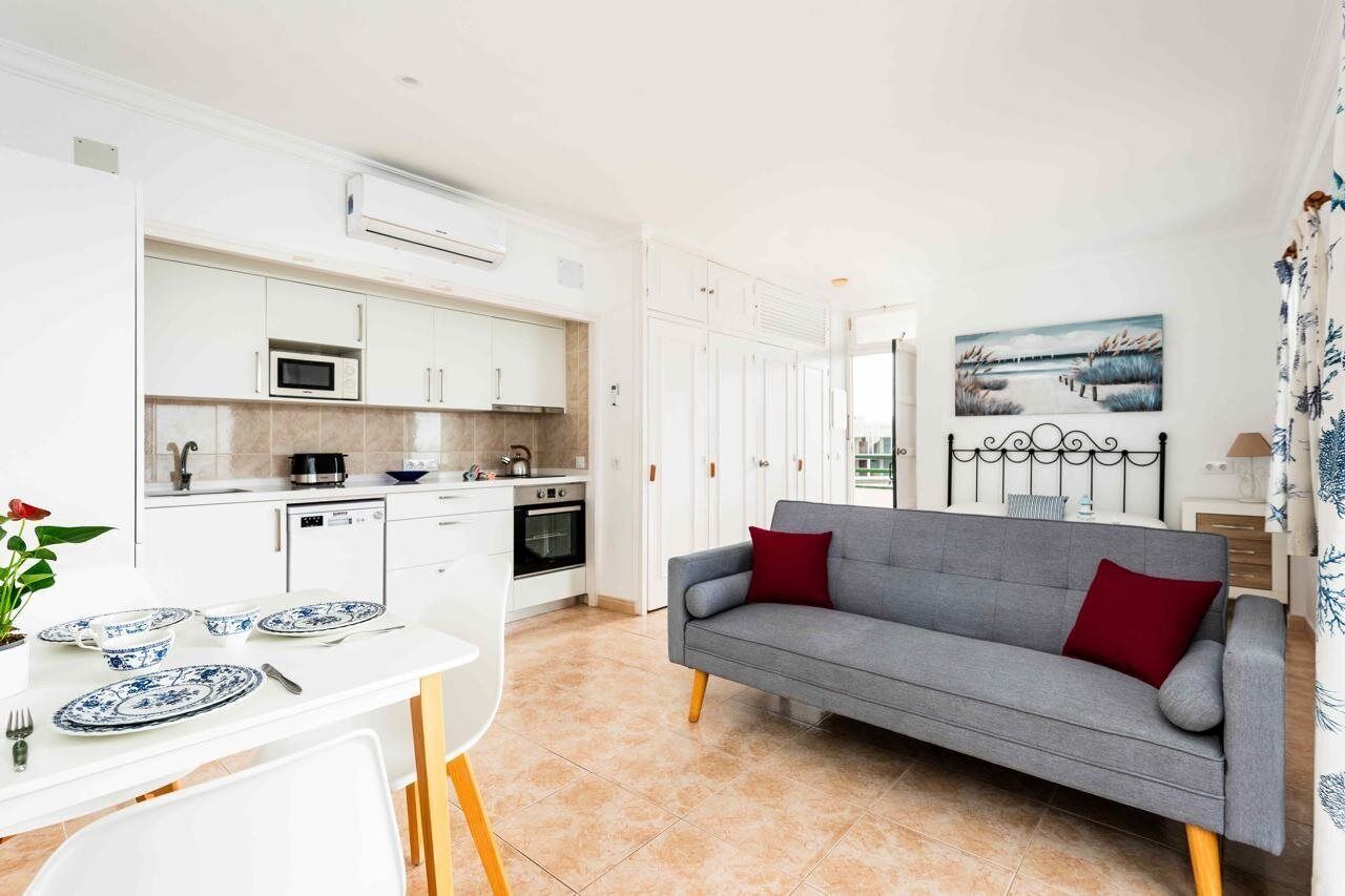 Una cocina moderna con armarios blancos, un microondas y una estufa. En la sala de estar, hay un sofá gris con cojines rojos.