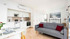 A modern kitchen with white cabinets, a microwave, and a stove. There is a grey sofa with red cushions in the living area. - Vistamar Apartments (Es Migjorn Gran)