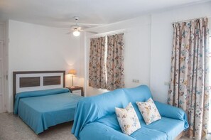 Un dormitorio con dos camas, un sofá azul, cortinas estampadas, un ventilador de techo y una lámpara de noche.