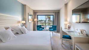 A hotel room with a large bed, two chairs, a desk, and a view of a pool and palm trees. - Barcelo Costa Ballena Golf & Spa (Rota)
