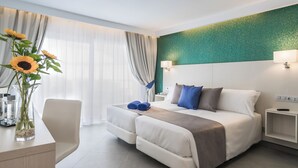 Ein modernes Hotelzimmer mit einem Bett, Nachttischen, einem Schreibtisch mit einer Vase Sonnenblumen und Blick ins Freie durch leichte Vorhänge.