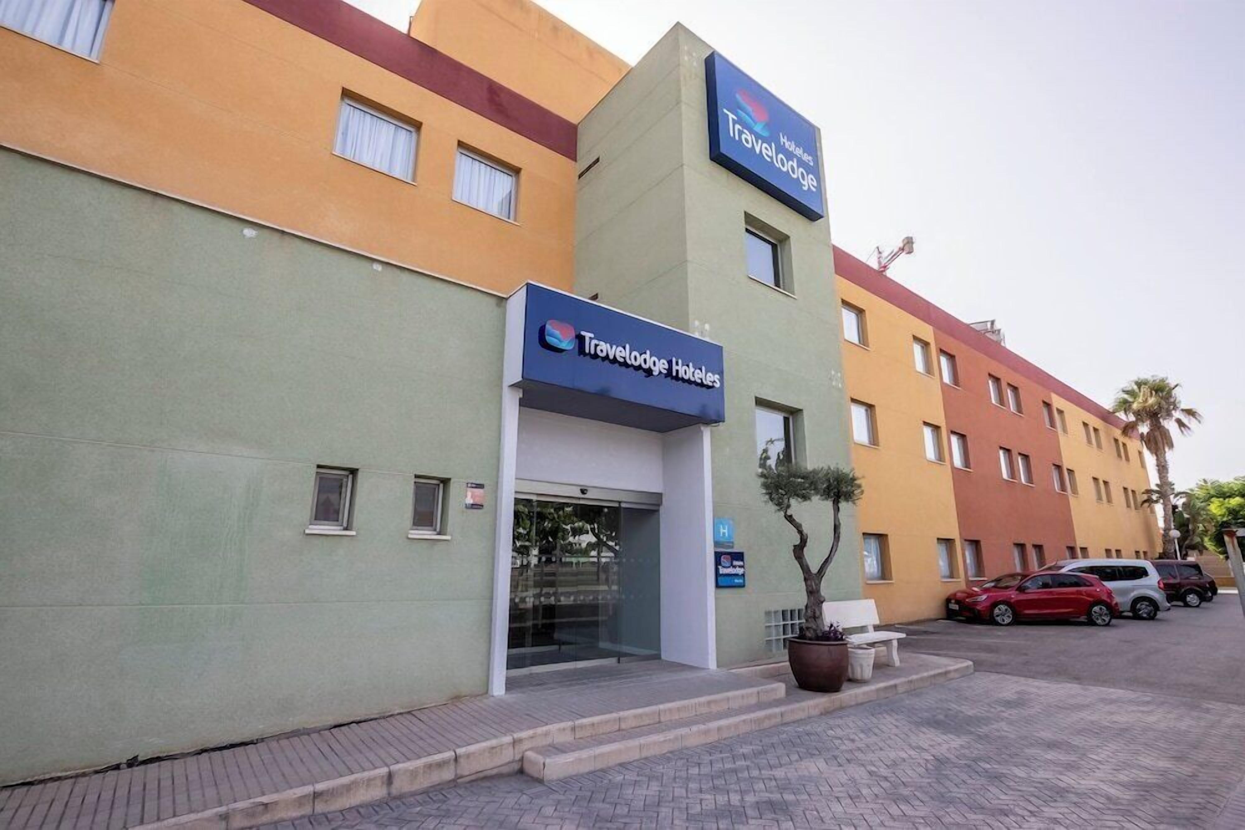 Un hotel Travelodge con fachada moderna, una zona de estacionamiento y un pequeño jardín con un banco.