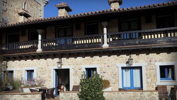 A stone building with a balcony, windows with blue shutters, and a small tree in the foreground.