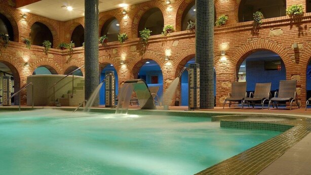 An indoor swimming pool with a brick wall, arched windows, and seating areas.