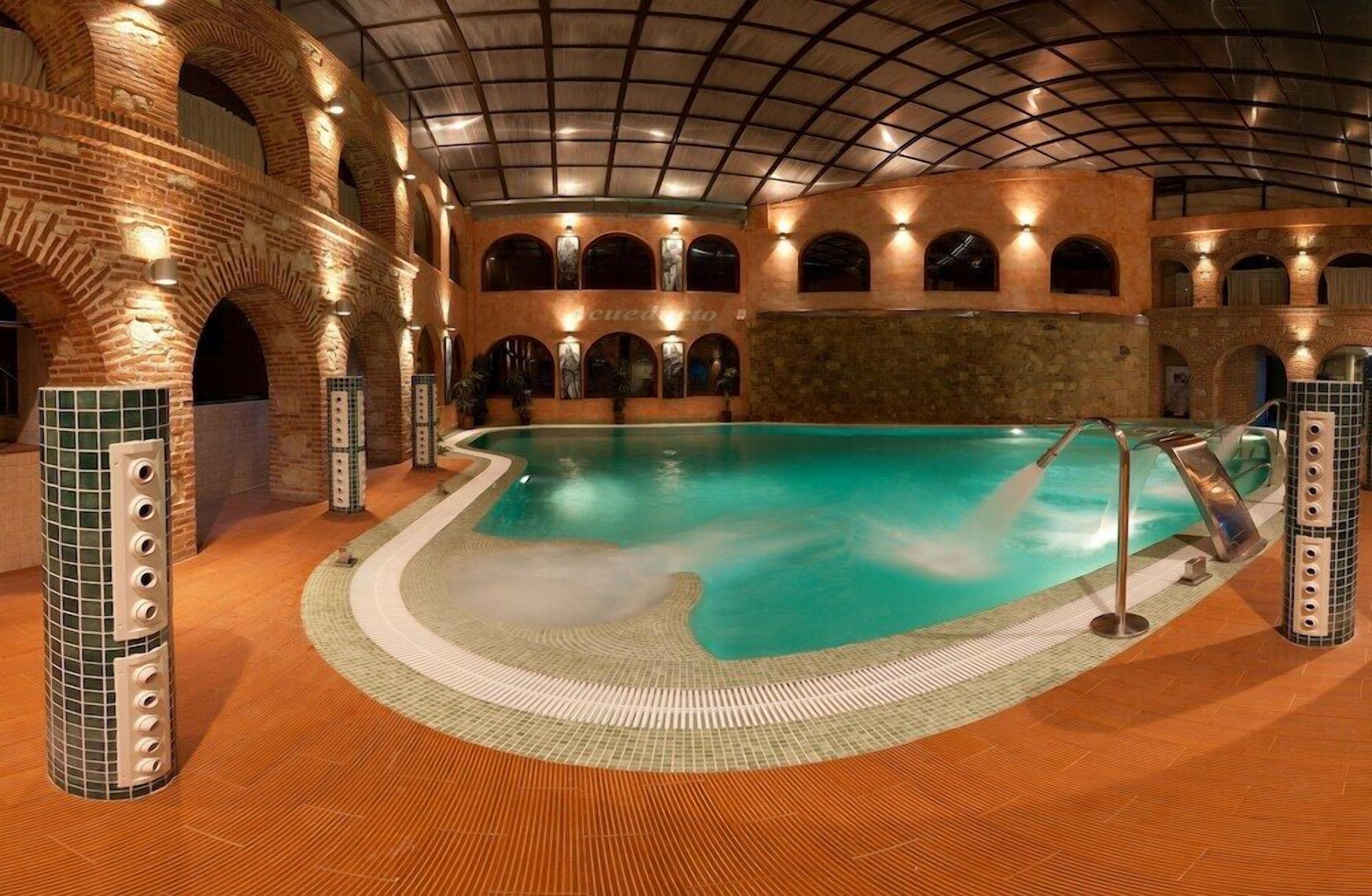 Una piscina interior con diseño curvo, rodeada de muros y arcos de ladrillo, y un suelo de baldosas.