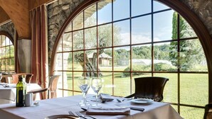 A dining table set with plates, cutlery, and wine glasses, with a view of a grassy landscape through large arched windows. - Hotel & Spa Monasterio de Boltana (Boltana)