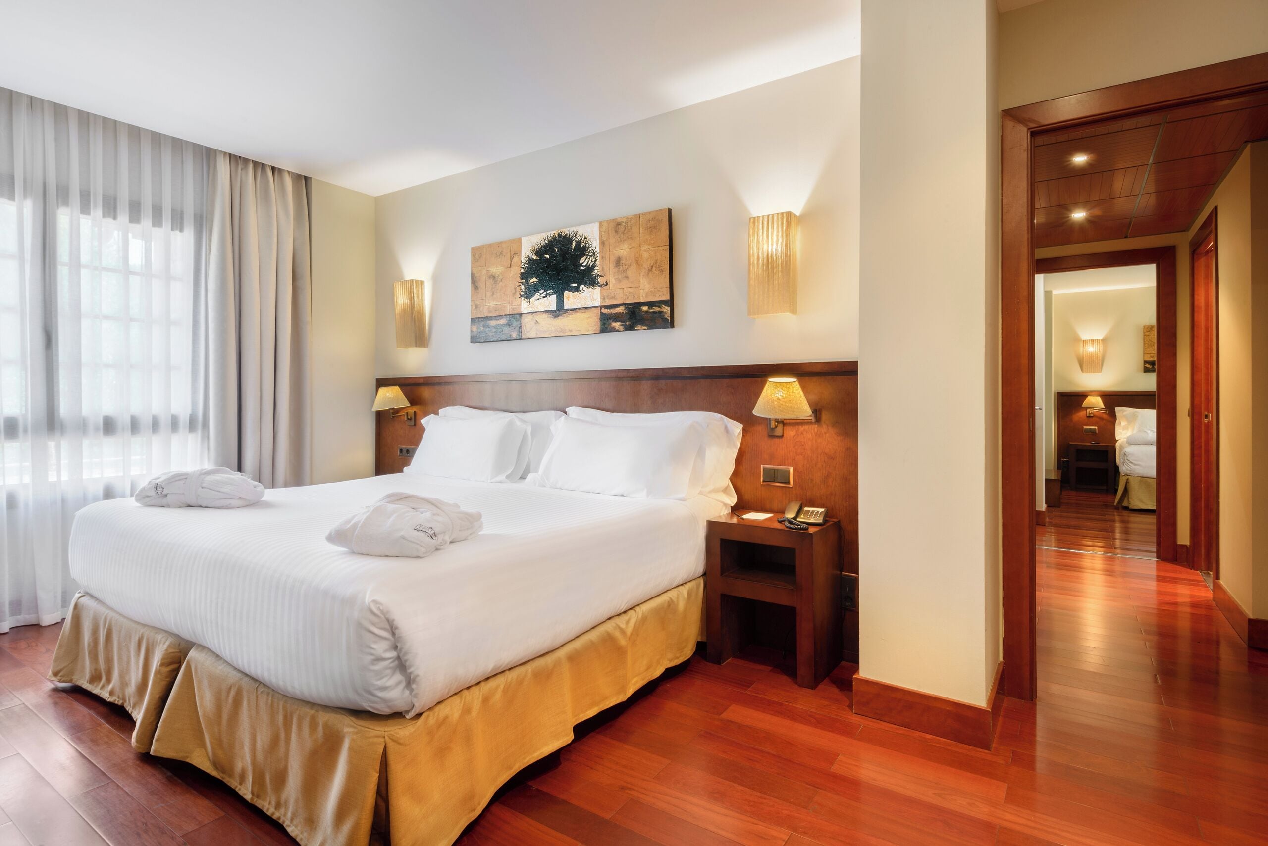 Habitación de hotel con una cama grande, cabecero de madera, dos mesitas de noche con lámparas, un cuadro en la pared y suelo de madera.
