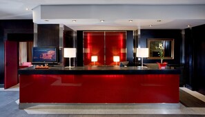 A modern hotel lobby with a red reception desk, two wall-mounted lamps, a television displaying 'MGM DIAMONDS', and a framed picture on the wall. - Eurostars Royal Tanau (Naut Aran)