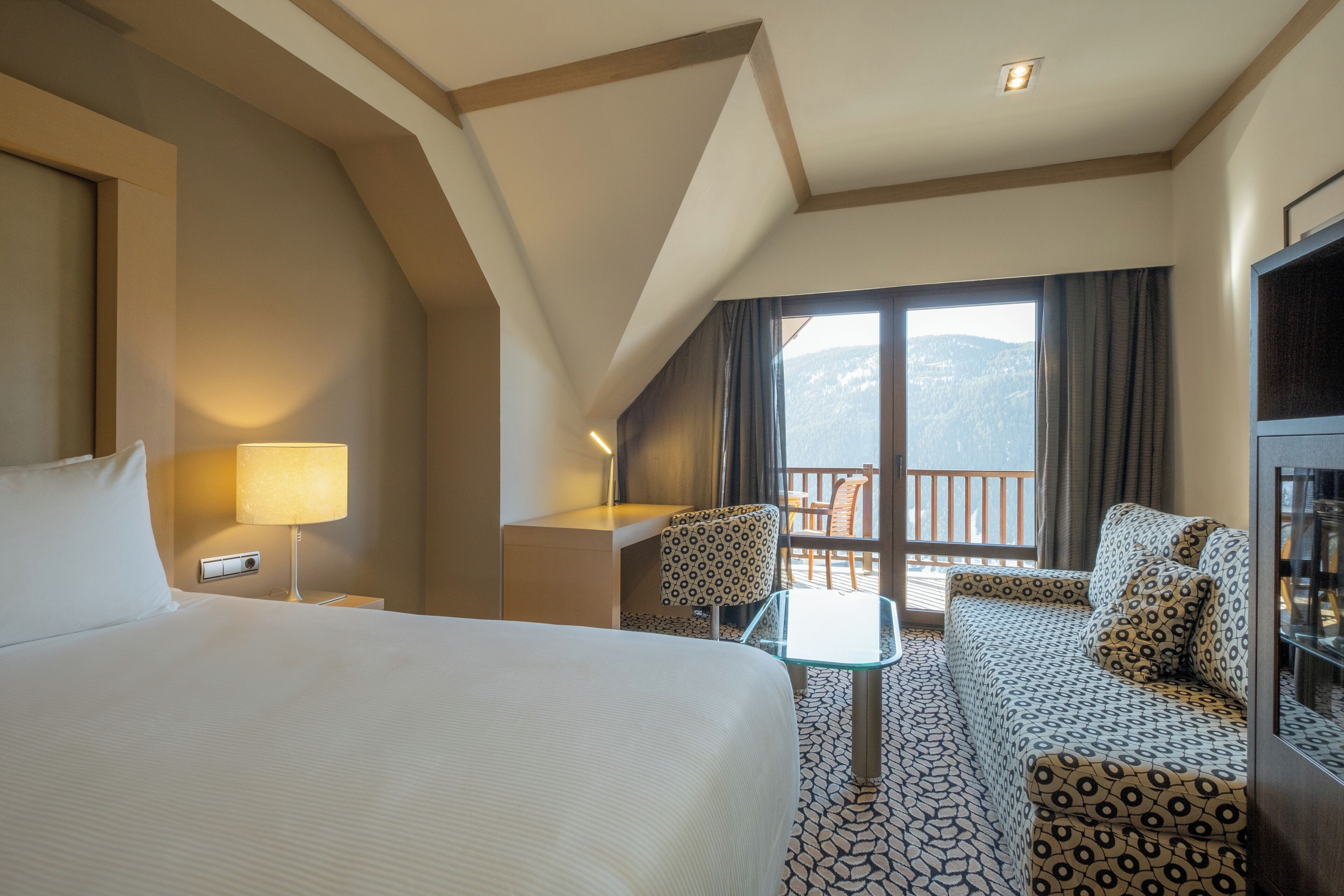 A hotel room with a large bed, a small table, a sofa, and a balcony with a mountain view.