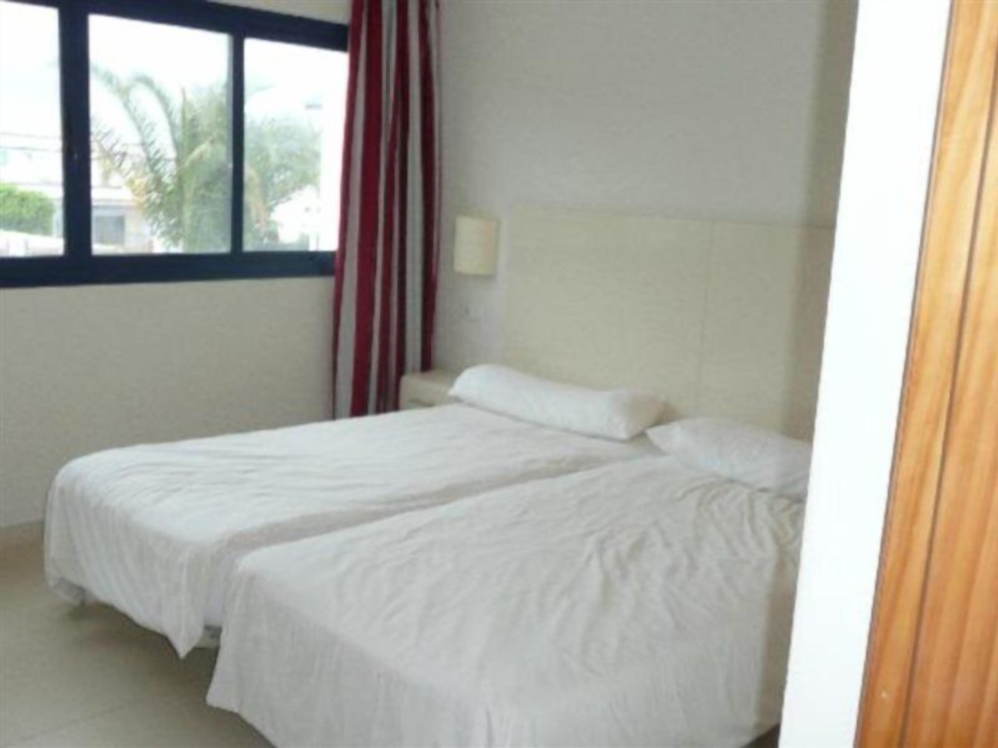 A hotel room with two single beds, a window with red curtains, and a view of palm trees outside.