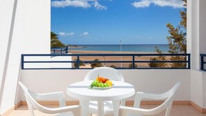 Un balcon avec une table et des chaises blanches, une assiette de fruits et une vue sur la plage et l’océan.