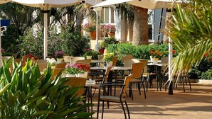Un patio al aire libre con mesas y sillas de mimbre, rodeado de plantas en macetas y sombrillas.