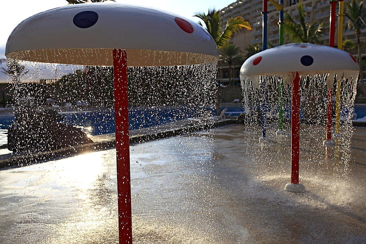 Two water fountains with red poles and white tops, surrounded by a sandy area with palm trees in the background.
