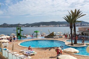 A pool area with lounge chairs, umbrellas, and a view of the sea and cityscape. - Playa Bella Beach Apartments (Sant Josep de sa Talaia)