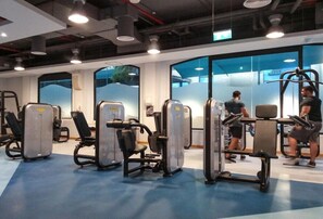Une salle de sport équipée de divers appareils d’exercice, dotée de grandes fenêtres et d’un sol bleu.