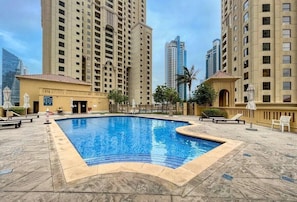 A swimming pool surrounded by high-rise buildings, with outdoor seating and a clear sky. - Ocean Views / Karaoke / Foosball / Gym / Pool (Dubai)