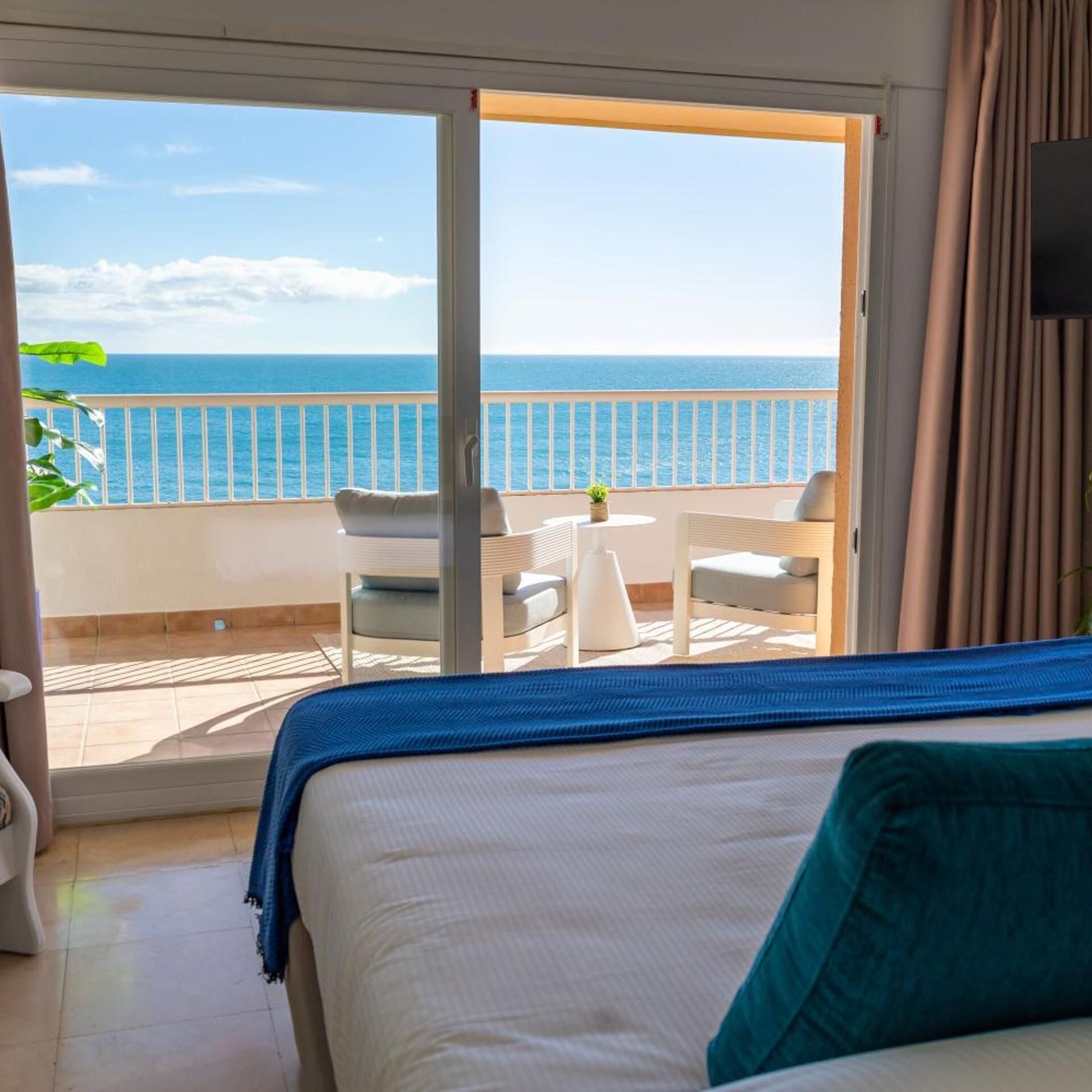 Une chambre d’hôtel avec un lit, une petite table et une chaise. La chambre offre une vue sur la mer grâce à une porte-fenêtre coulissante.
