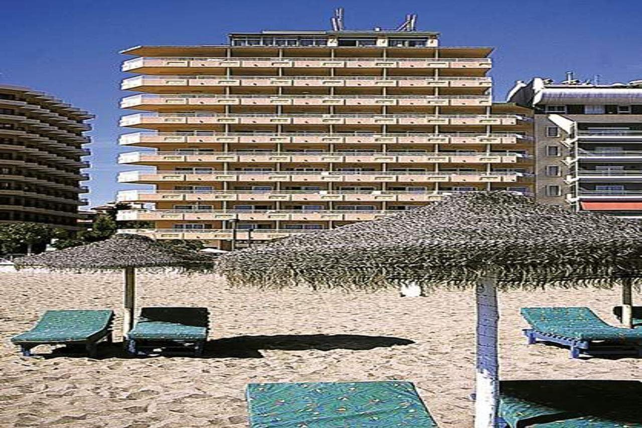 Un hotel de varios pisos con balcones, una zona de playa con tumbonas y sombrillas, y un cielo azul despejado.