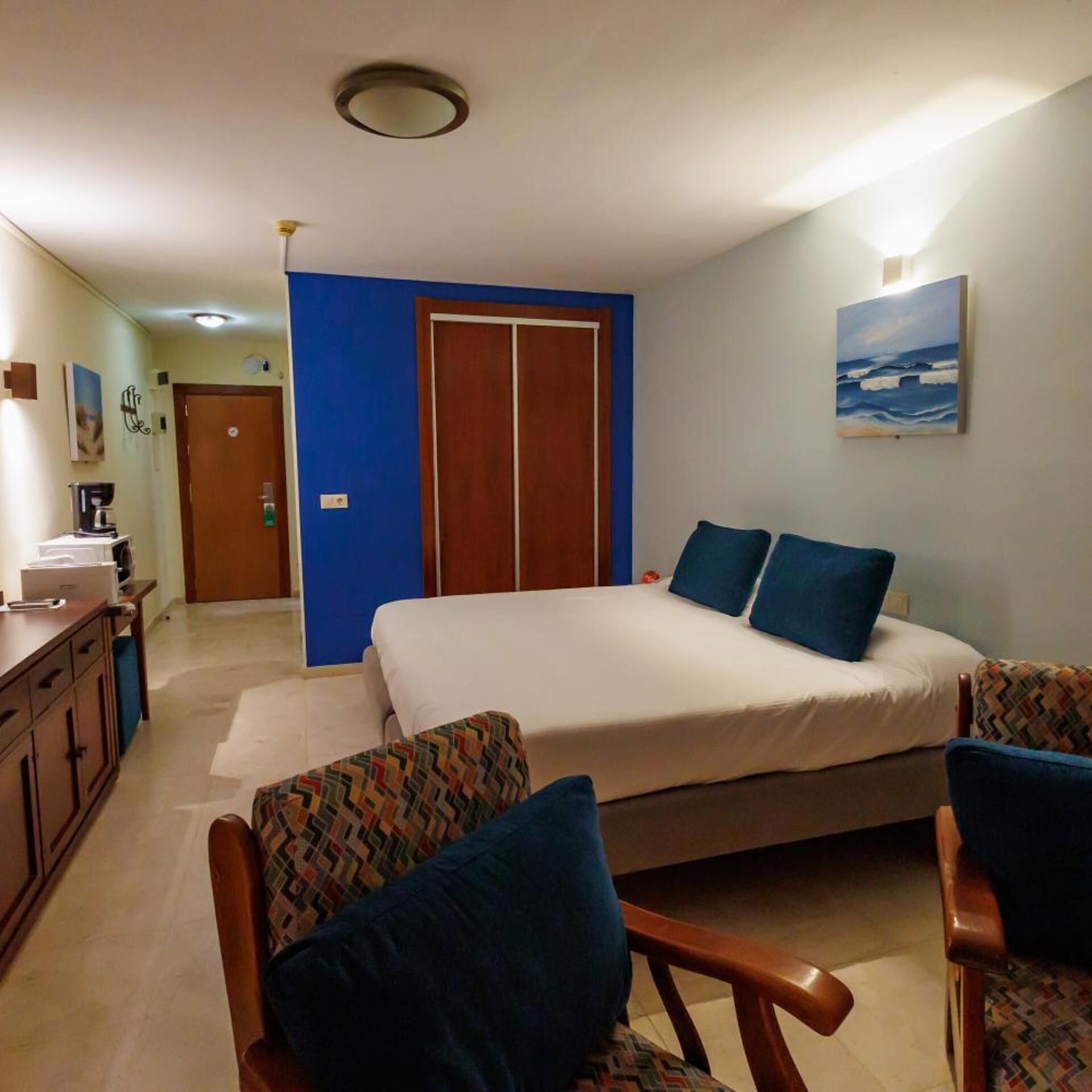 A hotel room with a bed, two chairs, a desk, and a painting on the wall.