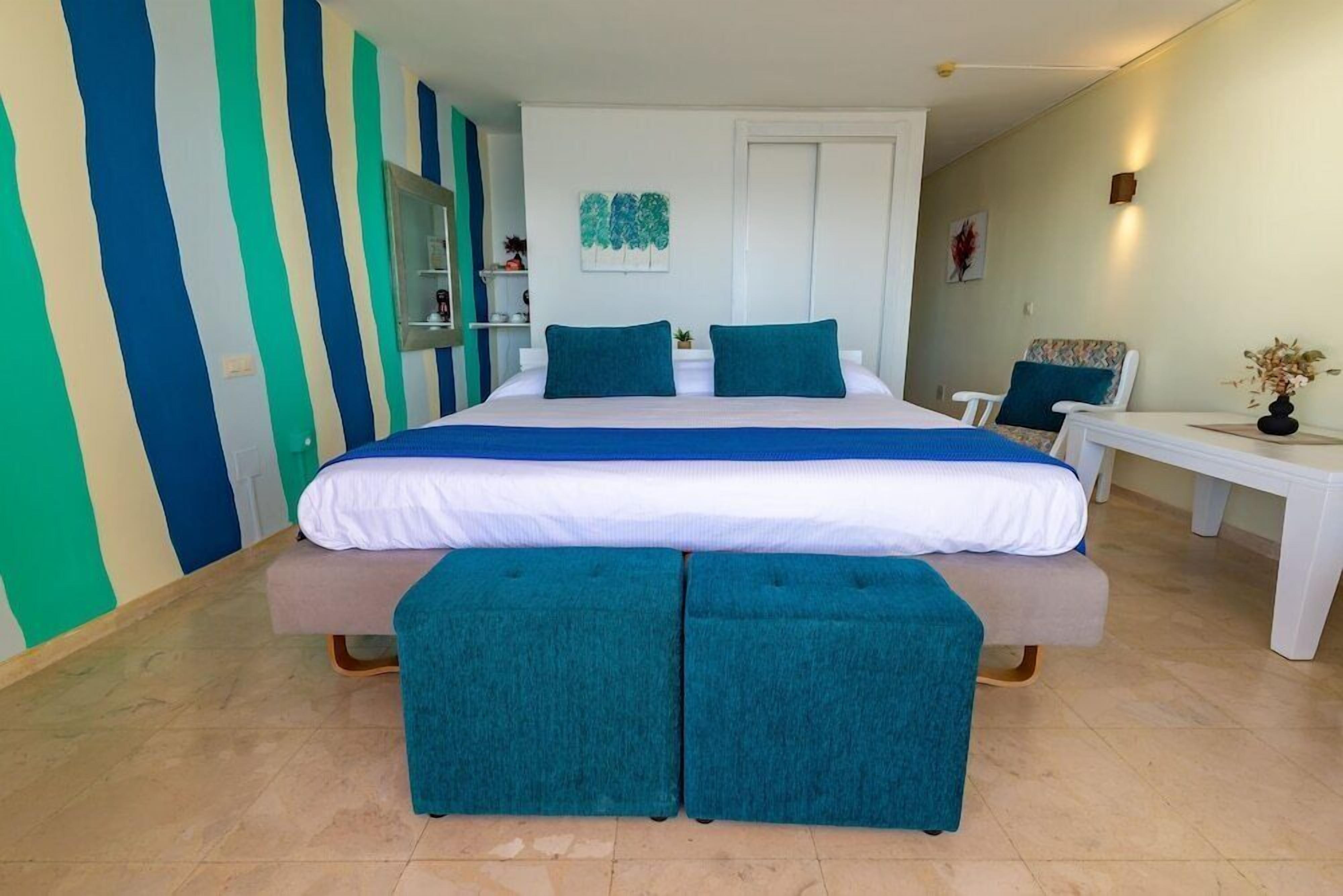 Een slaapkamer met een bed, twee kussens, een bankje, een stoel, een bijzettafeltje, een spiegel en een muur met verticale strepen.