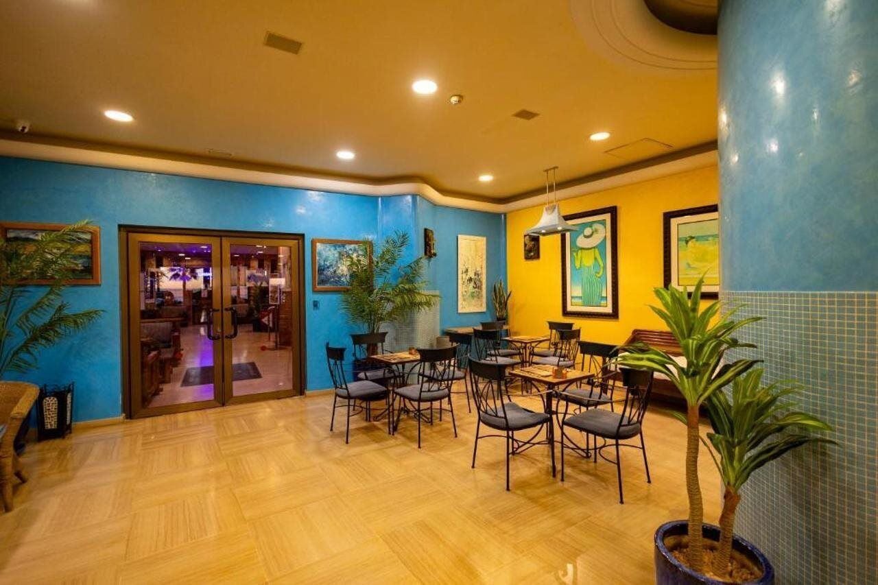 A brightly lit indoor space with a wooden floor, several tables and chairs, and potted plants.