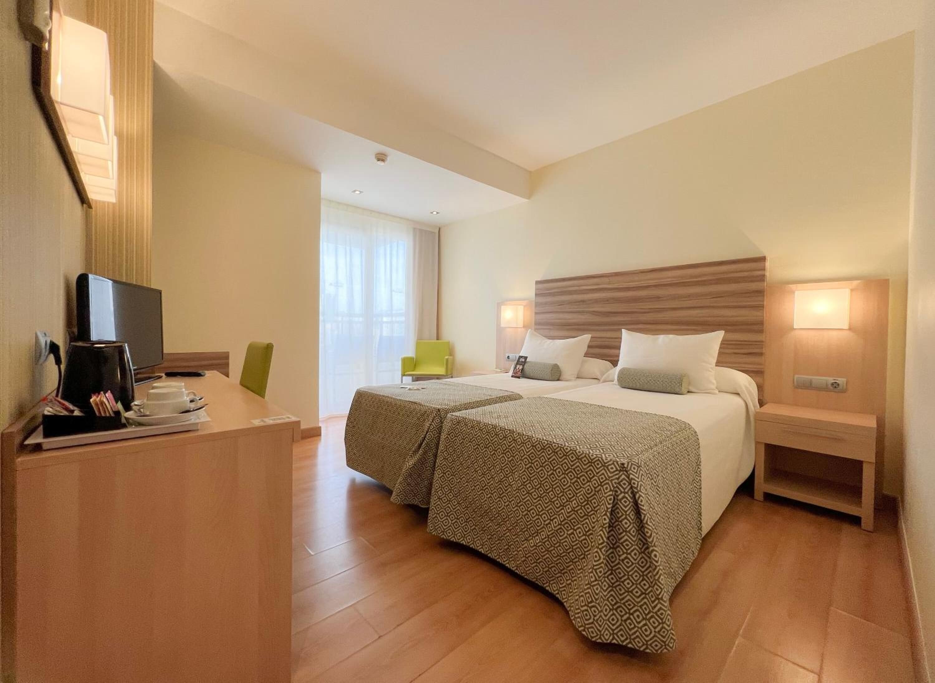 A hotel room with two beds, a desk with a coffee maker, a TV, and a window with curtains.