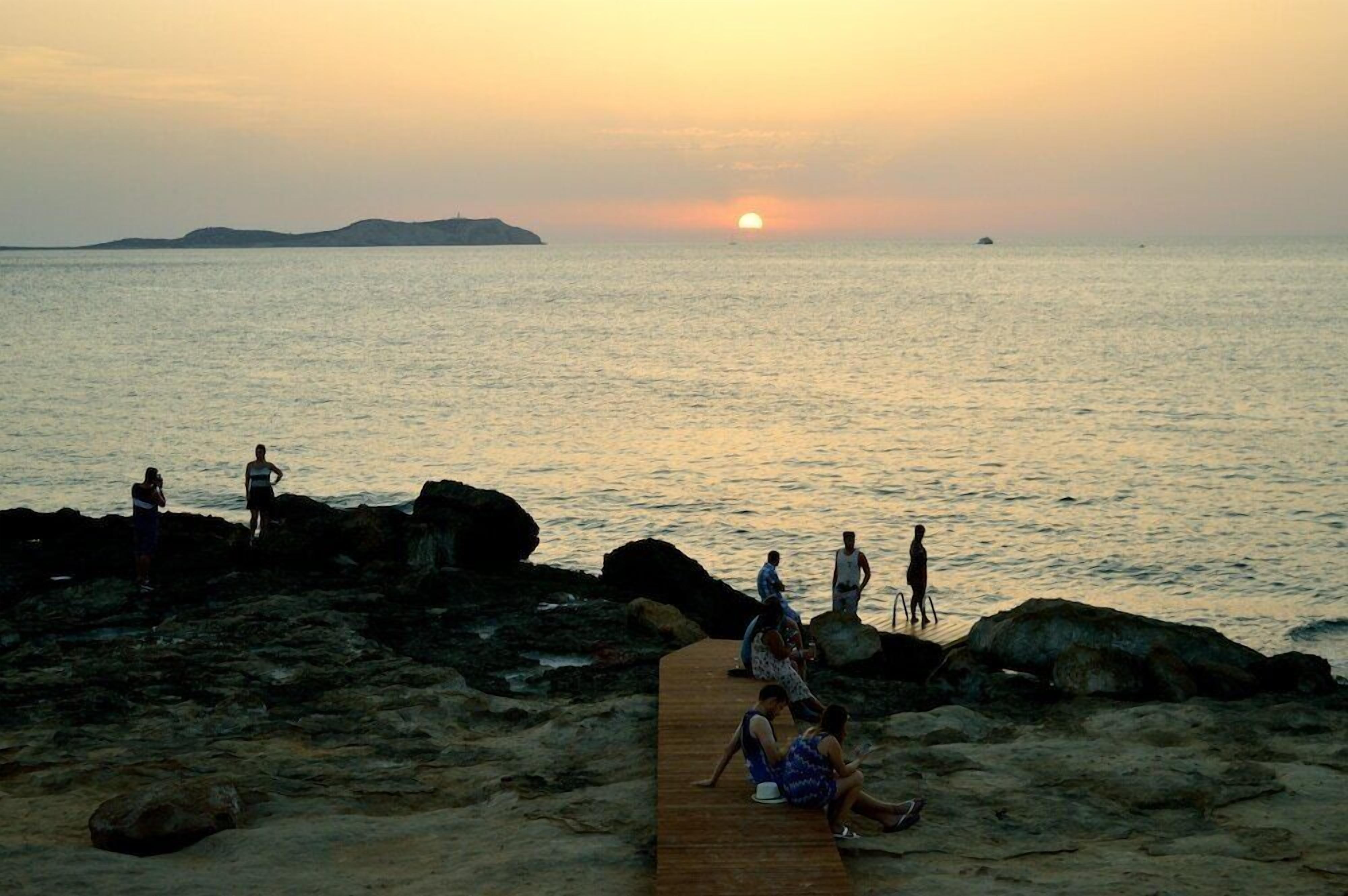 Une vue sur le coucher de soleil au-dessus d’une plage rocheuse, avec des personnes assises et debout sur une jetée en bois.