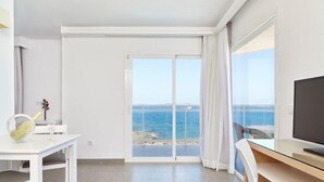 A room with a large window offering a view of the sea, a flat-screen TV, a dining table with chairs, and a small table with a bottle and glass. - Aparthotel Nereida (Sant Josep de sa Talaia)