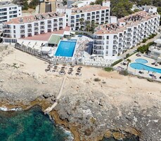 Un hotel costero con varios balcones, una piscina central y un litoral rocoso.