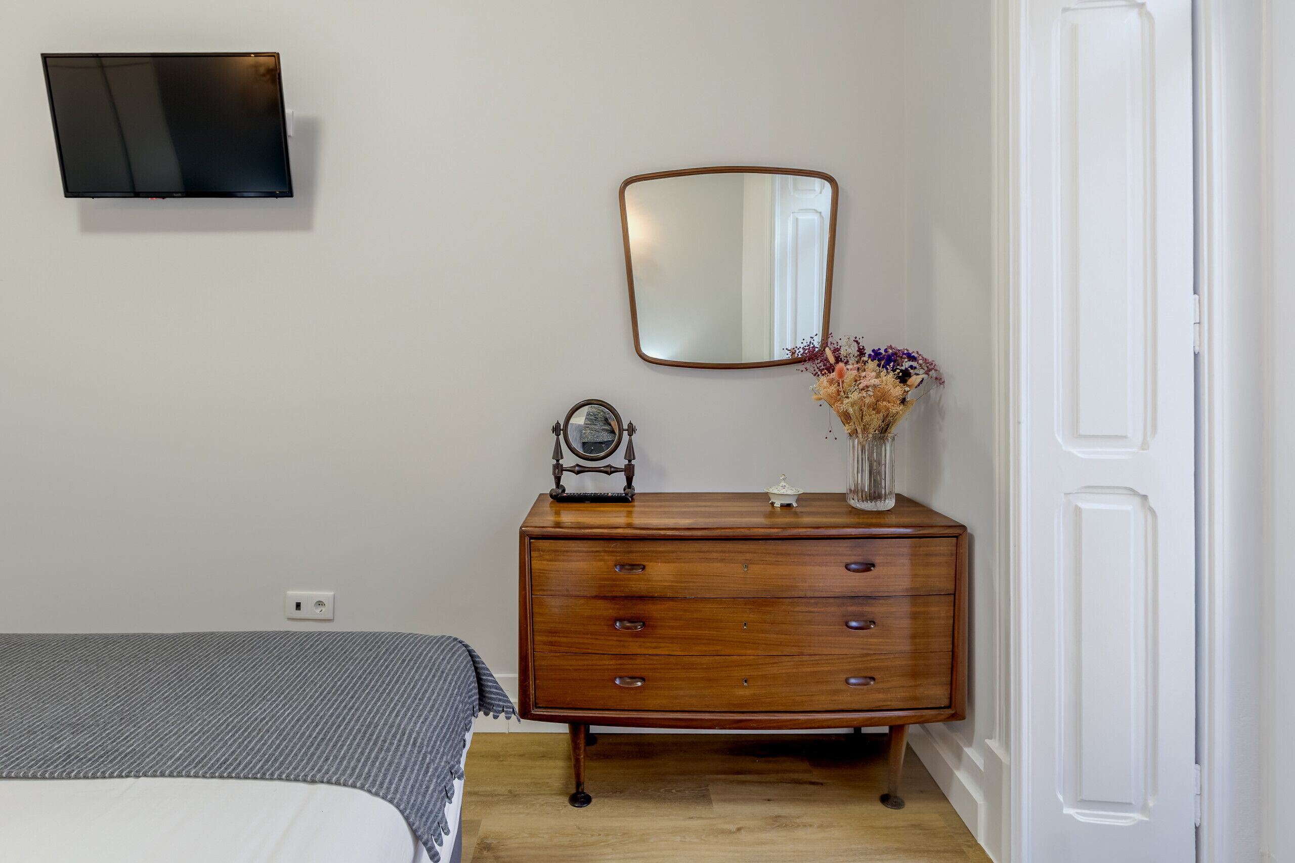A bedroom with a bed, a dresser, a mirror, and a wall-mounted TV.