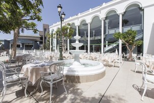 An outdoor dining area with white tables and chairs, a central fountain, and a building with large windows and arches in the background.