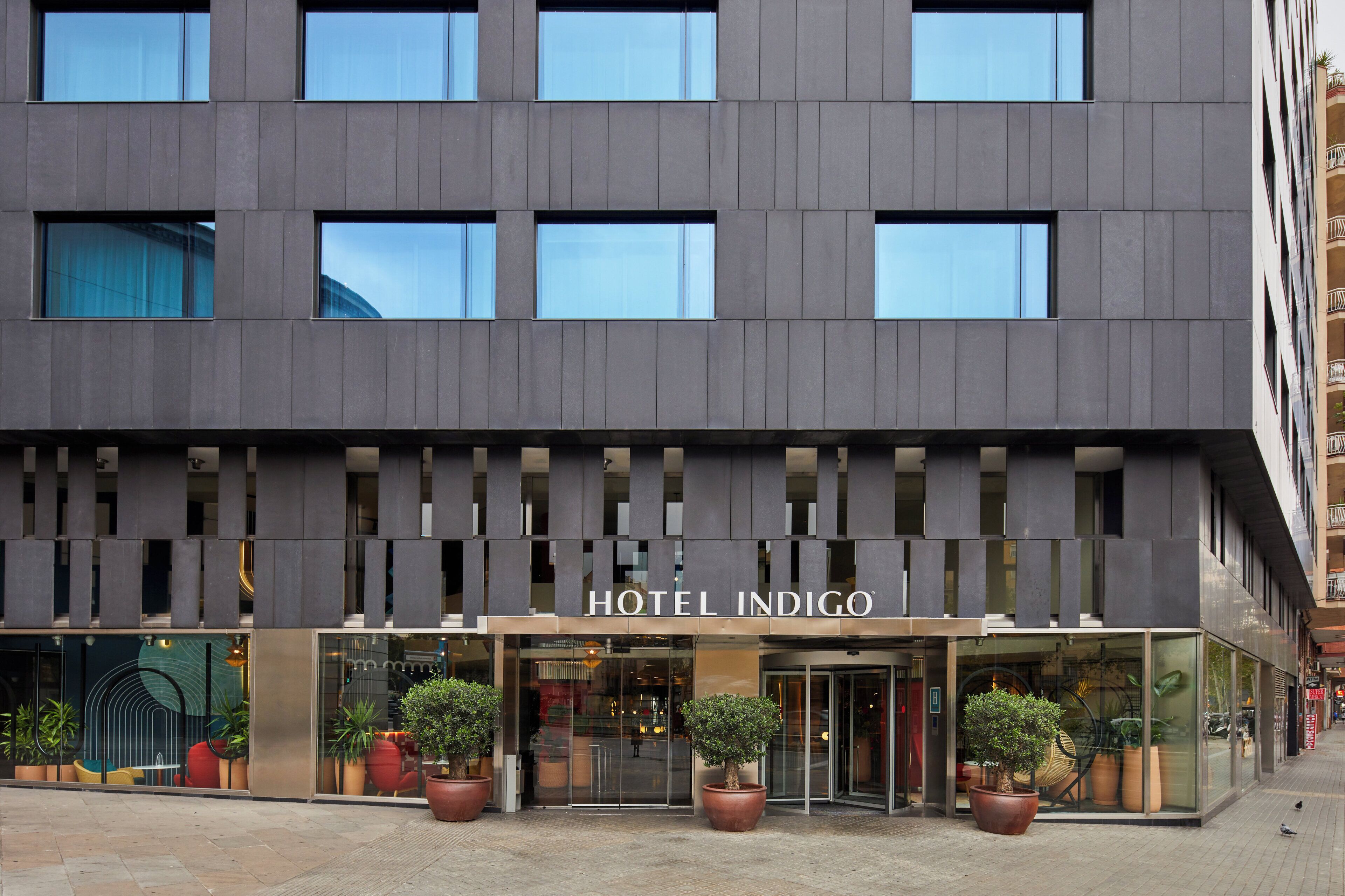 Un hôtel moderne avec une façade en verre, de grandes plantes en pot et un ciel dégagé.