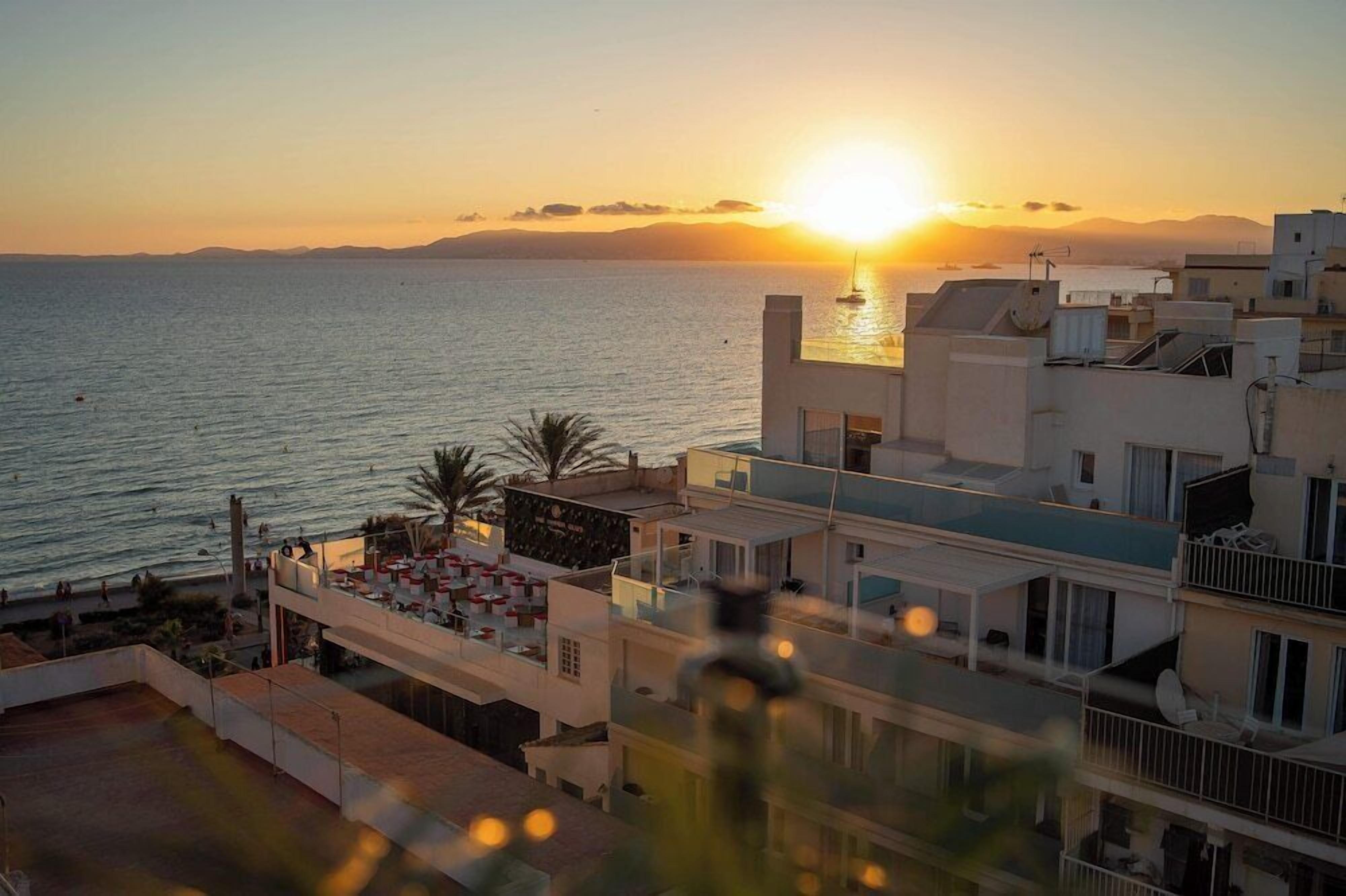 A coastal view with a sunset over the sea, modern buildings, and a rooftop bar with outdoor seating.