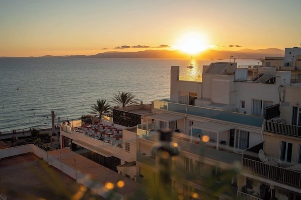 Vista costera con un atardecer sobre el mar, edificios modernos y un bar en la azotea con mesas al aire libre.